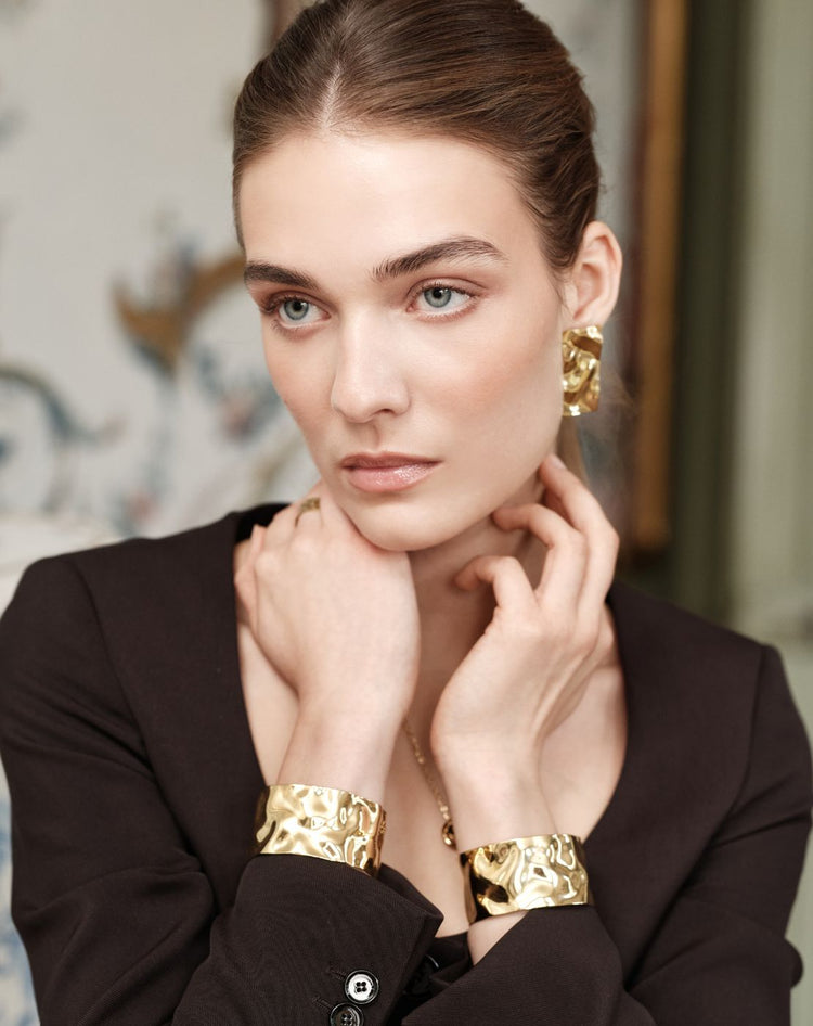 Woman wearing the bold Noem earrings and Noem bangles in gold from Avelore with a blurred background