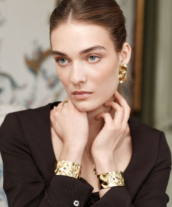 Woman wearing the bold Noem earrings and Noem bangles in gold from Avelore with a blurred background