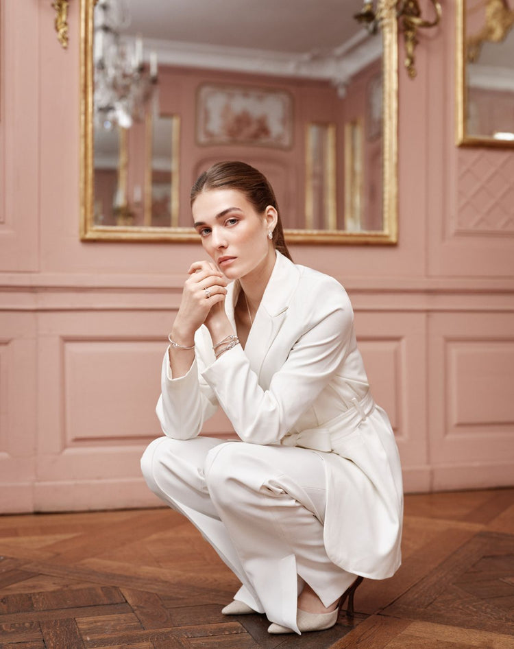 Woman wearing silver jewelry from Avelore and a white suit posing in a room with pink walls and gold accents.