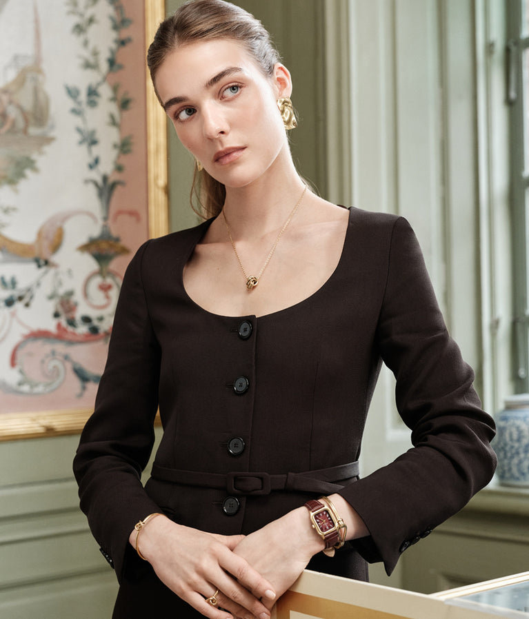 Woman standing by a piano in a room with decorative wall art wearing the bold Noem earrings paired with the square burgundy and gold leather watch Aima.