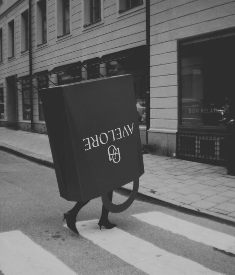 Woman walking in high heels in an oversized shopping bag with 'Avelore' logo on a city street.