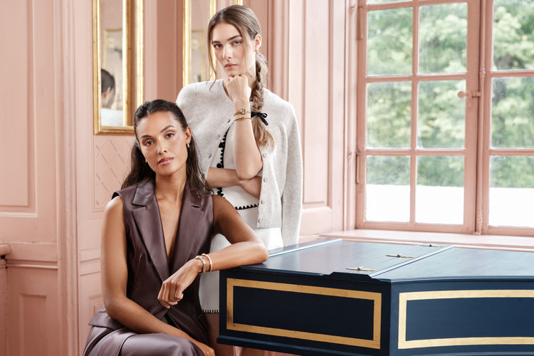 Two women wearing Avelore jewelry standing and sitting by a blue piano in a pink room.